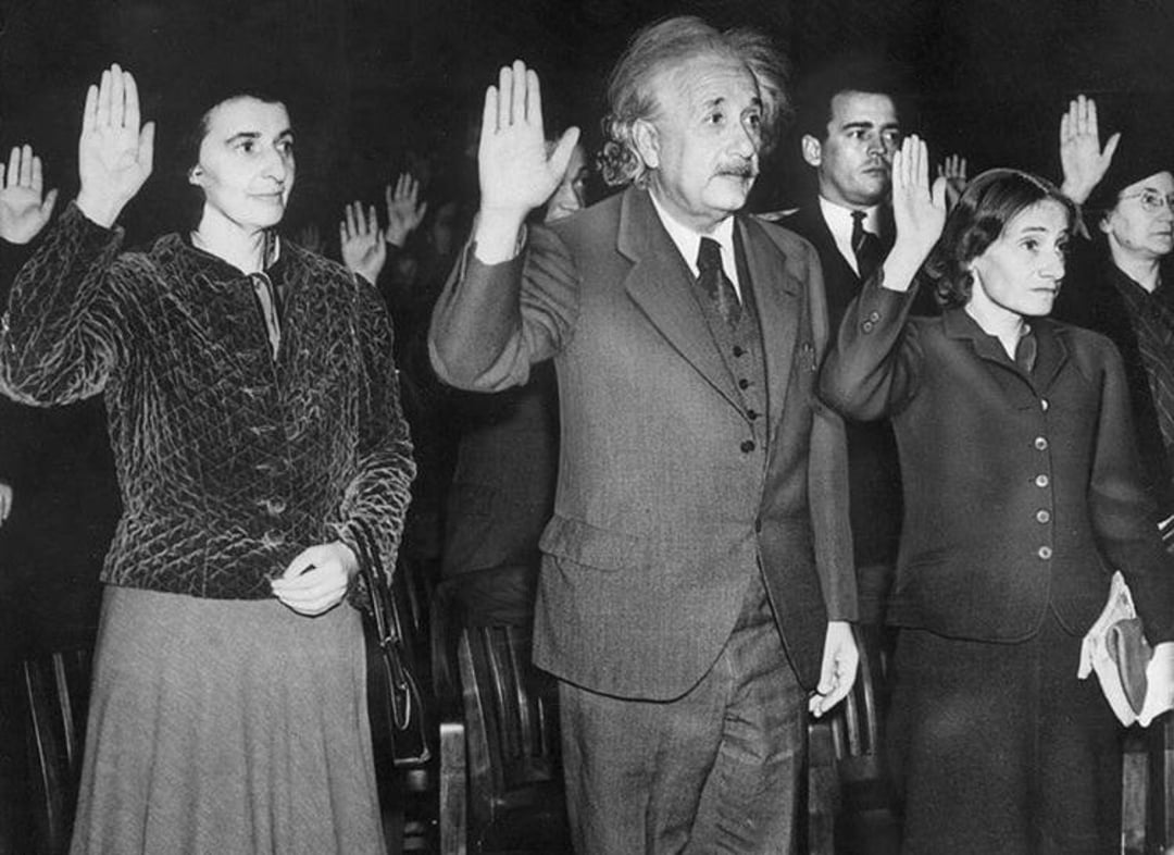 A group of people, including a man with wild hair and a mustache, stand with their right hands raised as if taking an oath. They are dressed in formal clothing in a black and white photo.