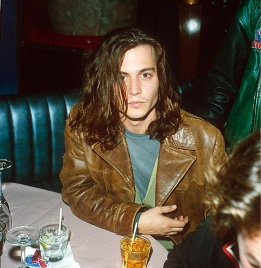 A person with long brown hair wearing a brown leather jacket sits at a table in a booth, next to two drinks. The background is dimly lit, and another person is partially visible in the foreground.