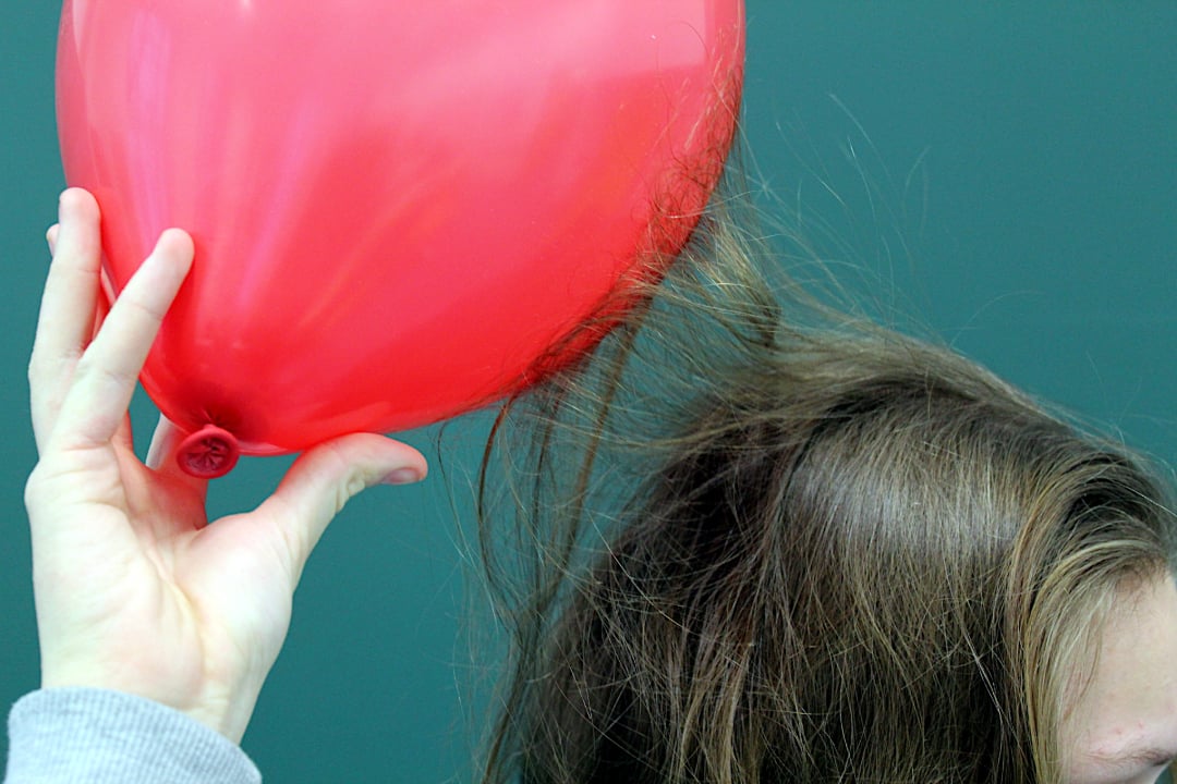 A hand holds a red balloon near a person's brown hair, causing the hair to stand up due to static electricity, with a plain greenish background.