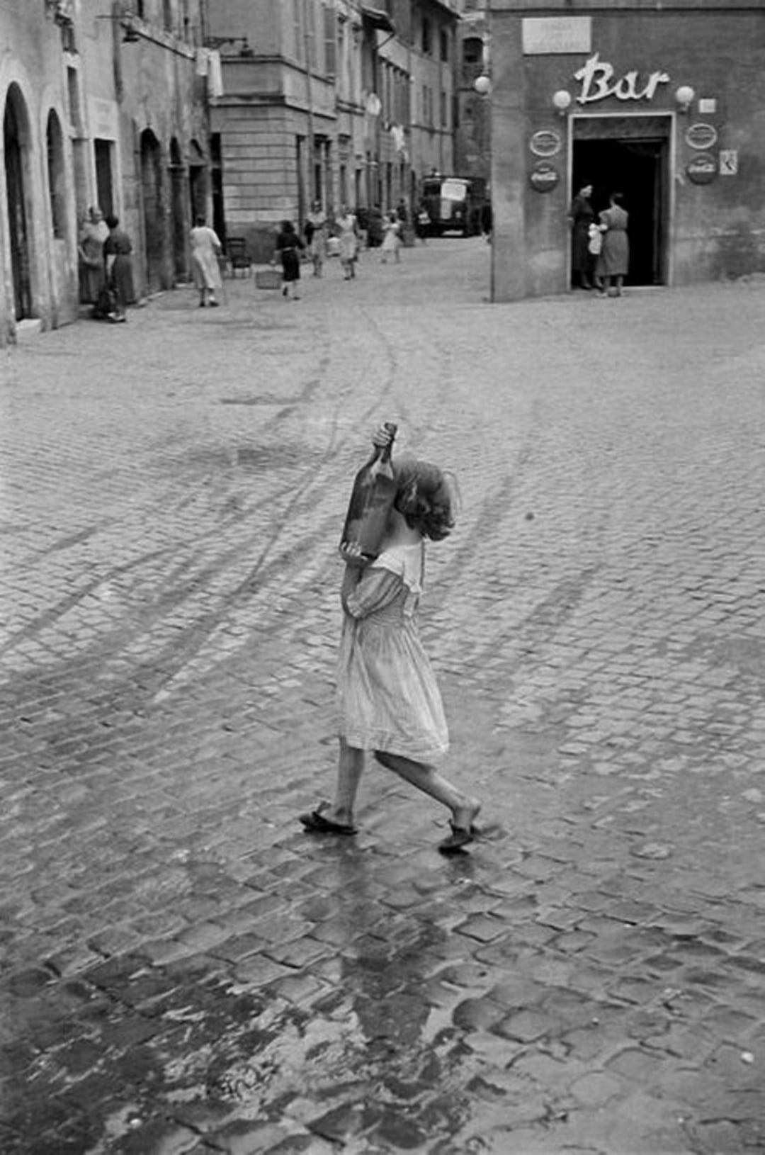 A young girl in a dress walks barefoot across a cobblestone street, carrying a large bottle on her shoulder. In the background, people stroll near old stone buildings and a bar with a sign above the entrance.