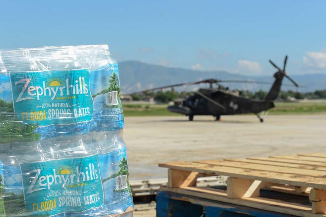 A close-up of Zephyrhills bottled water packs with a military helicopter and wooden pallets on an airstrip in the background. Mountains and a clear sky are visible in the distance.