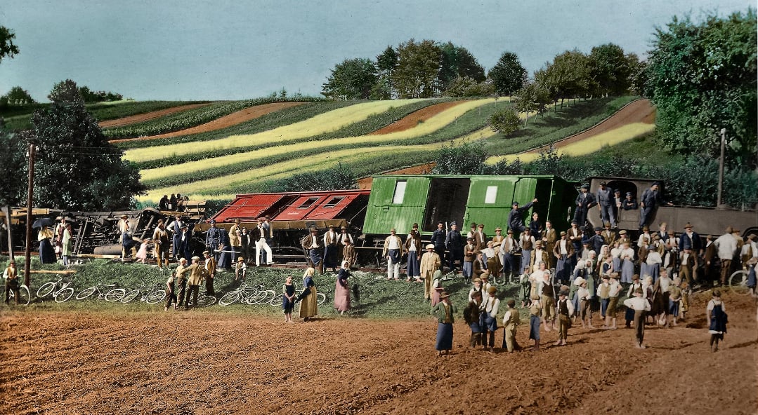 A large group of people, including adults and children, stand and walk near colorful train cars on a dirt field, with patterned farmland and trees on a hill in the background.