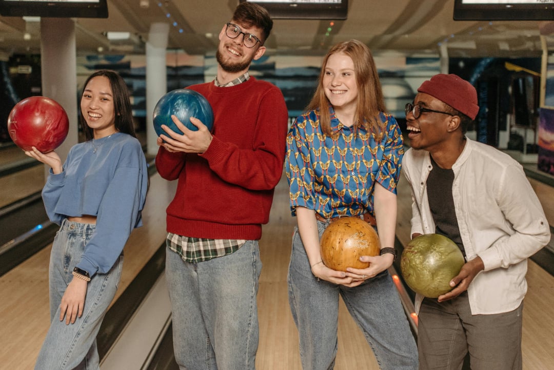 Four young adults stand in a bowling alley, smiling and holding colorful bowling balls. They appear happy and ready to play, with bowling lanes visible in the background.