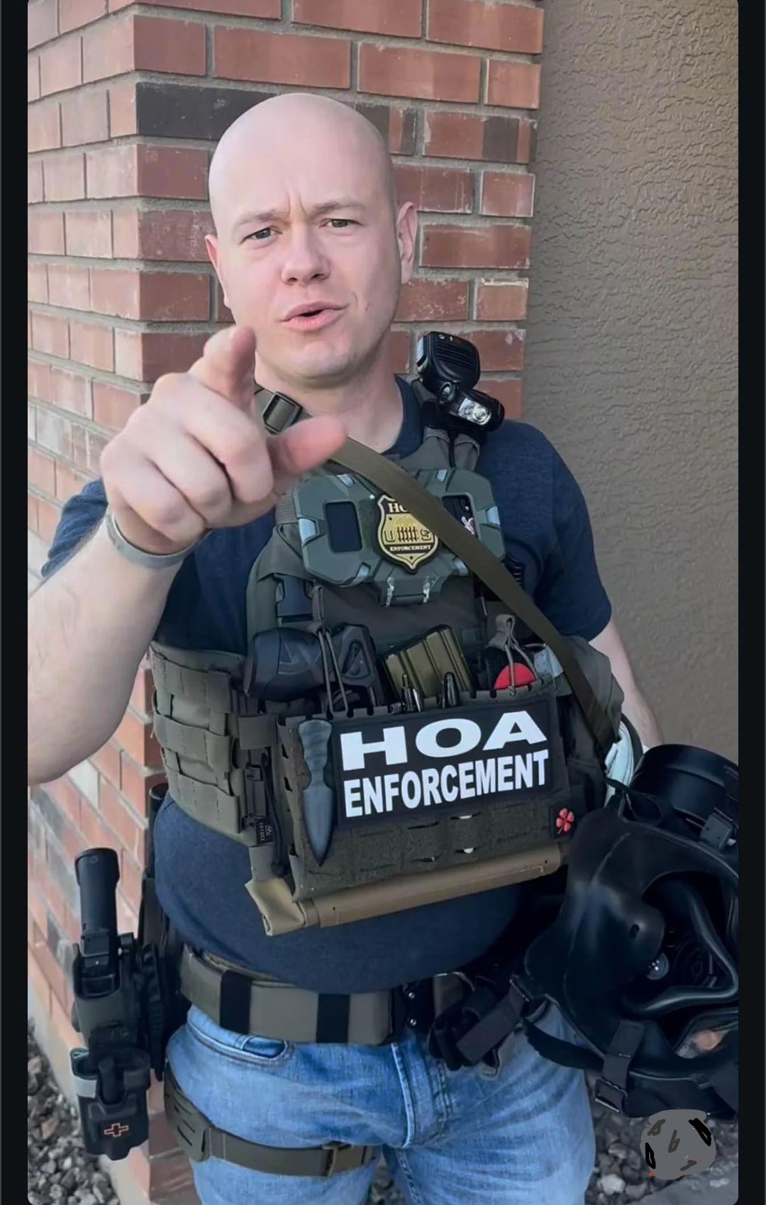 A bald man in tactical gear stands in front of a brick wall, pointing towards the camera. He is wearing a vest labeled “HOA Enforcement” and holding a black helmet in his left hand.