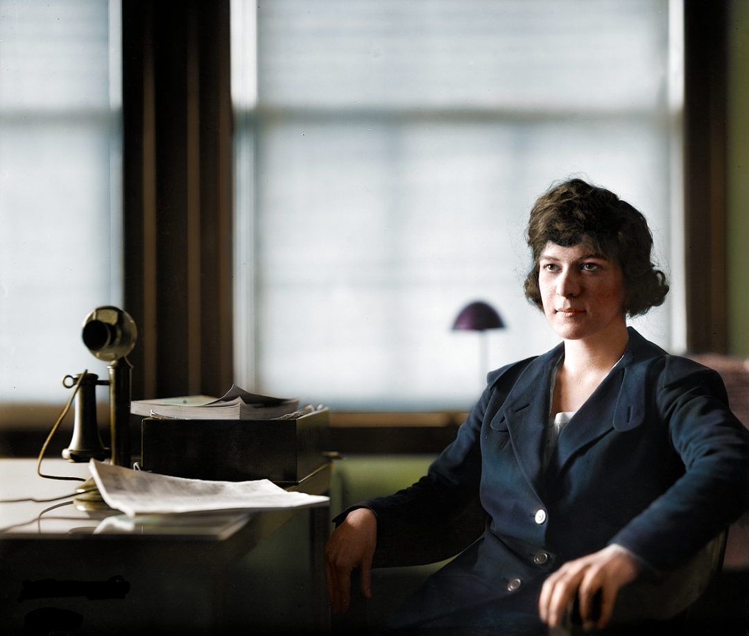 A woman in a dark suit sits at a desk with papers, a telephone, and a box. She looks at the camera, and soft light streams through large windows behind her.