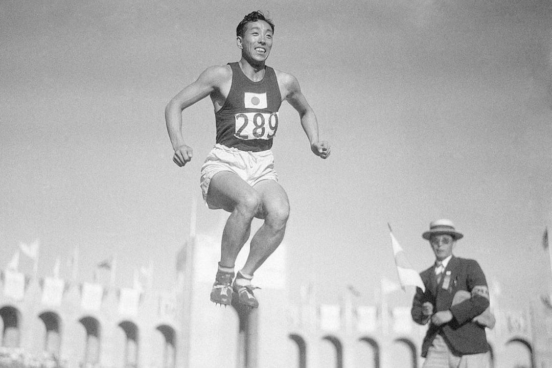 A male athlete with bib number 289 and a Japanese flag on his shirt is mid-air during a long jump event at a stadium. An official in a hat and blazer stands nearby holding a flag.