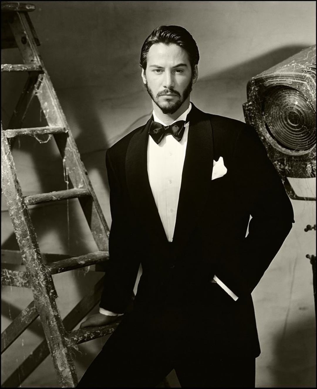 A man in a tuxedo with a bow tie stands confidently beside a weathered ladder and a large studio light, posing in a classic black-and-white portrait with a serious expression.