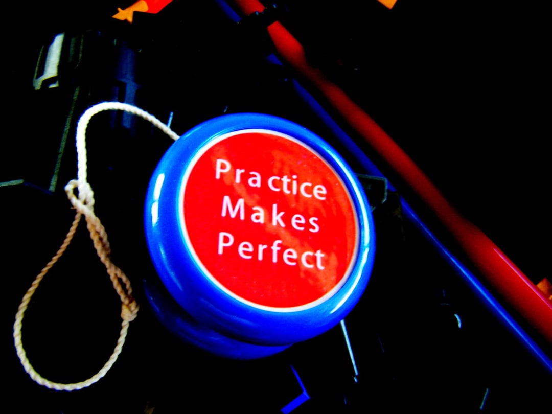 A blue yo-yo with a white string features a red center displaying the words "Practice Makes Perfect" in white text, set against a dark, blurred background.