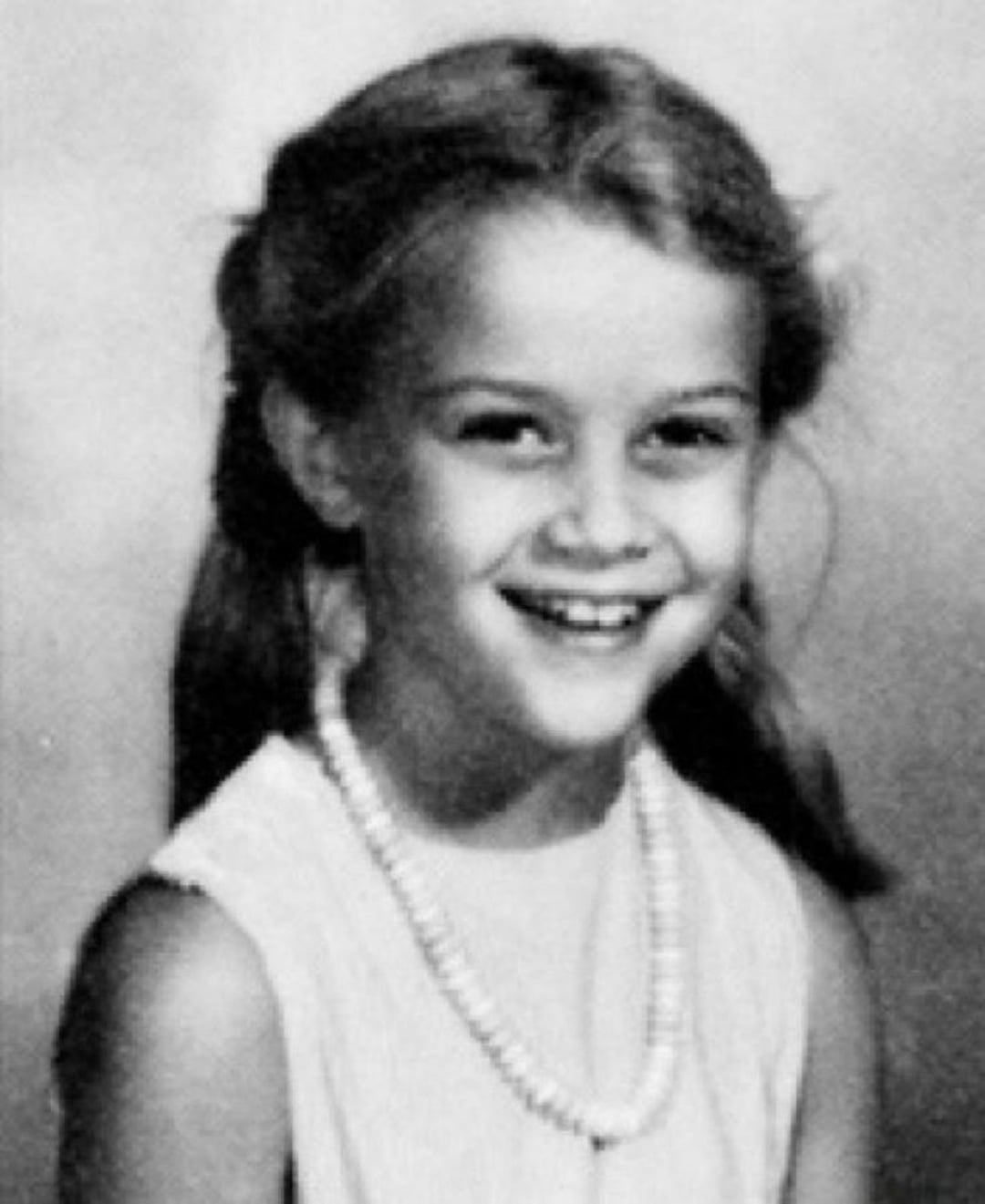 A young girl with long hair in pigtails smiles at the camera. She is wearing a sleeveless top and a beaded necklace. The photo is black and white and has a soft, vintage appearance.