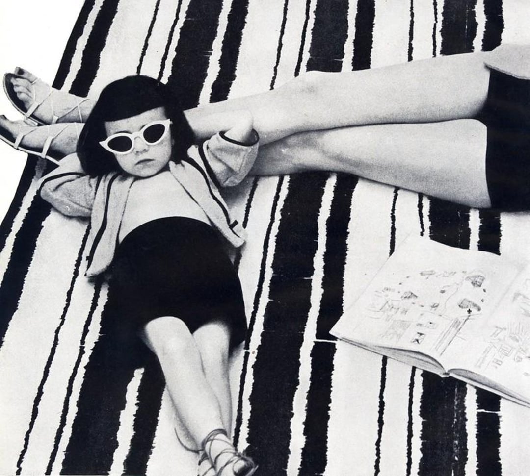 A young girl wearing sunglasses and sandals lounges on a striped rug next to an adult's crossed legs and an open illustrated book. The image is in black and white.