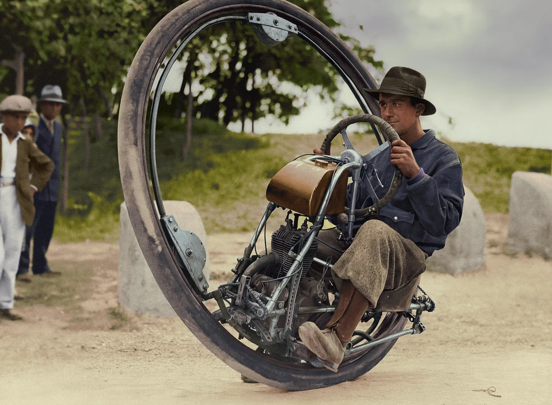 A man wearing a hat and jacket rides a large, single-wheeled monowheel motorcycle, while several people stand in the background observing him outdoors.