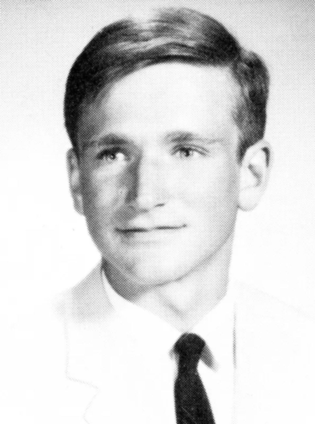 Black-and-white portrait of a young man with short, neatly combed hair, wearing a light-colored jacket, white shirt, and dark tie, looking slightly to the side and smiling softly.