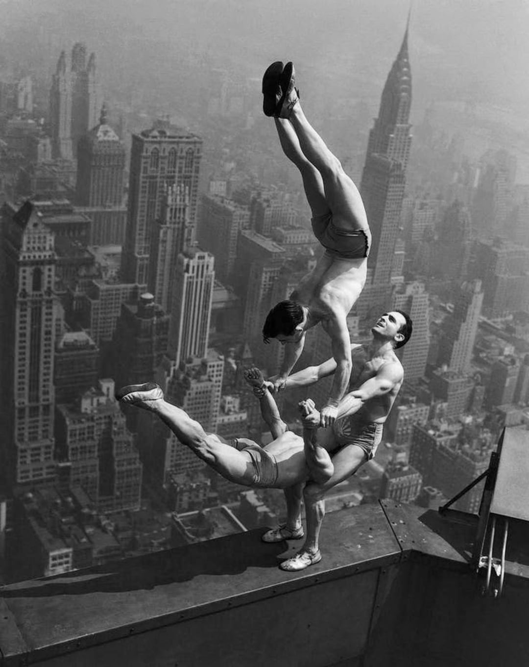 Three male acrobats perform a balancing act on the edge of a skyscraper rooftop, high above a cityscape with tall buildings and the Chrysler Building in the background.