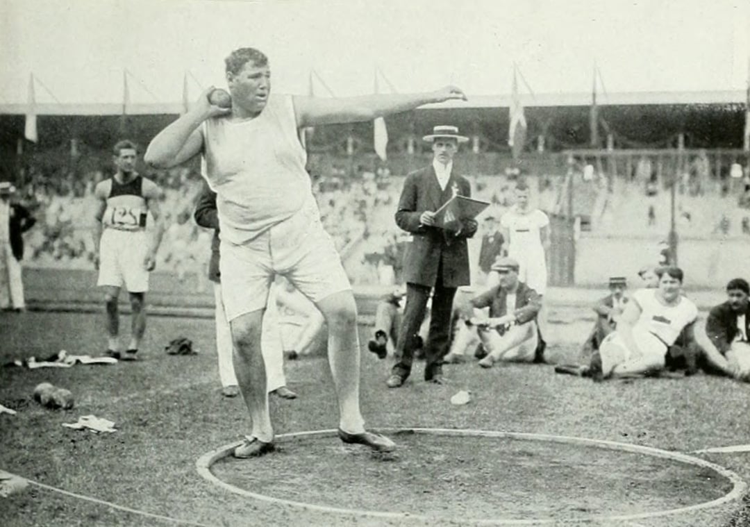 A male shot put athlete prepares to throw in a stadium during an old-fashioned sports event, with spectators, officials, and competitors in vintage attire watching in the background.