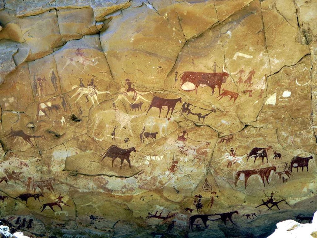 Ancient rock wall covered in prehistoric cave paintings depicting people, cattle, and animals in various scenes, using reddish-brown and white pigments. The rock surface is cracked and uneven.