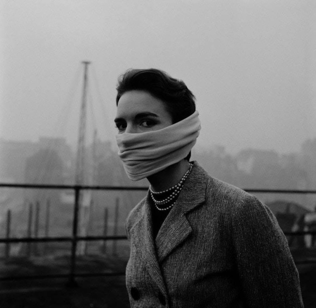 A woman in a coat and layered pearl necklaces stands outdoors on a foggy day, with a scarf wrapped around her lower face. A metal railing and blurred structures are visible in the background.