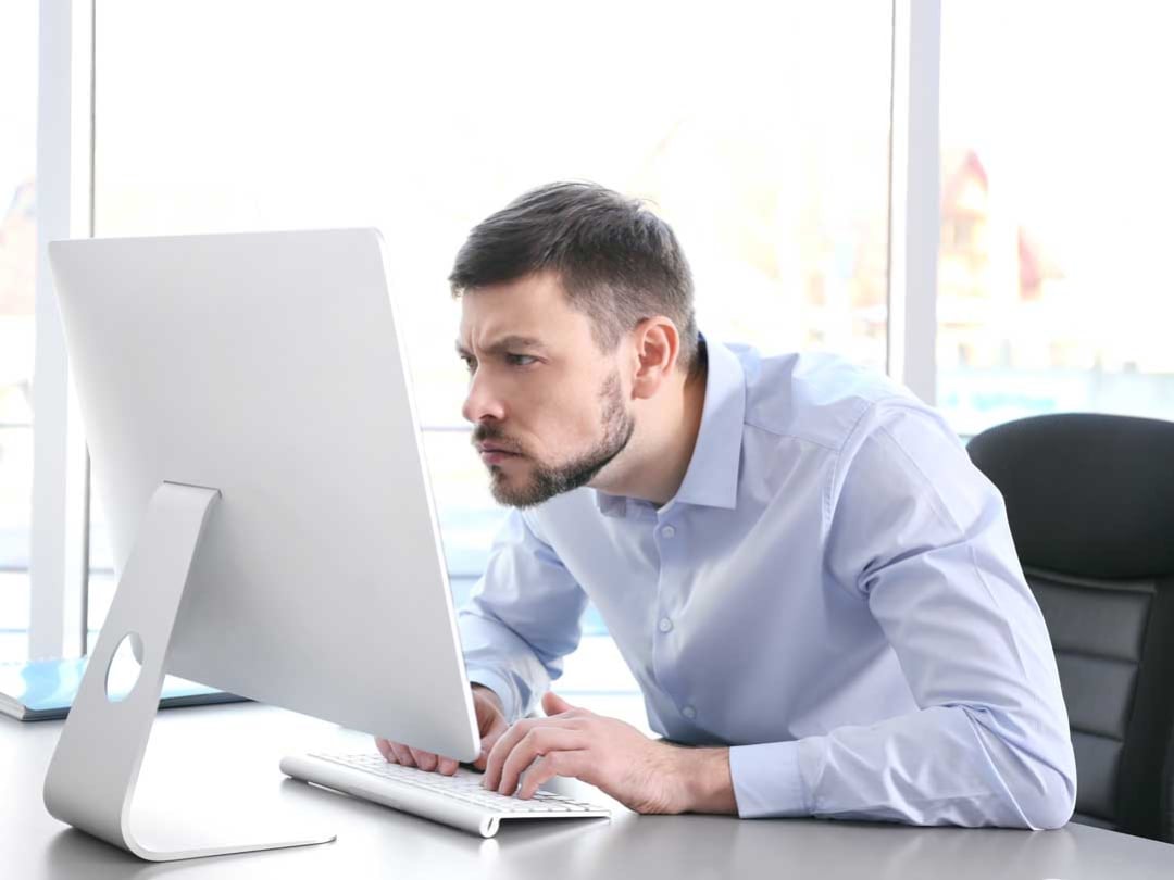 A man in a light blue shirt sits at a desk, leaning forward and squinting closely at a desktop computer monitor, appearing to have difficulty seeing the screen.