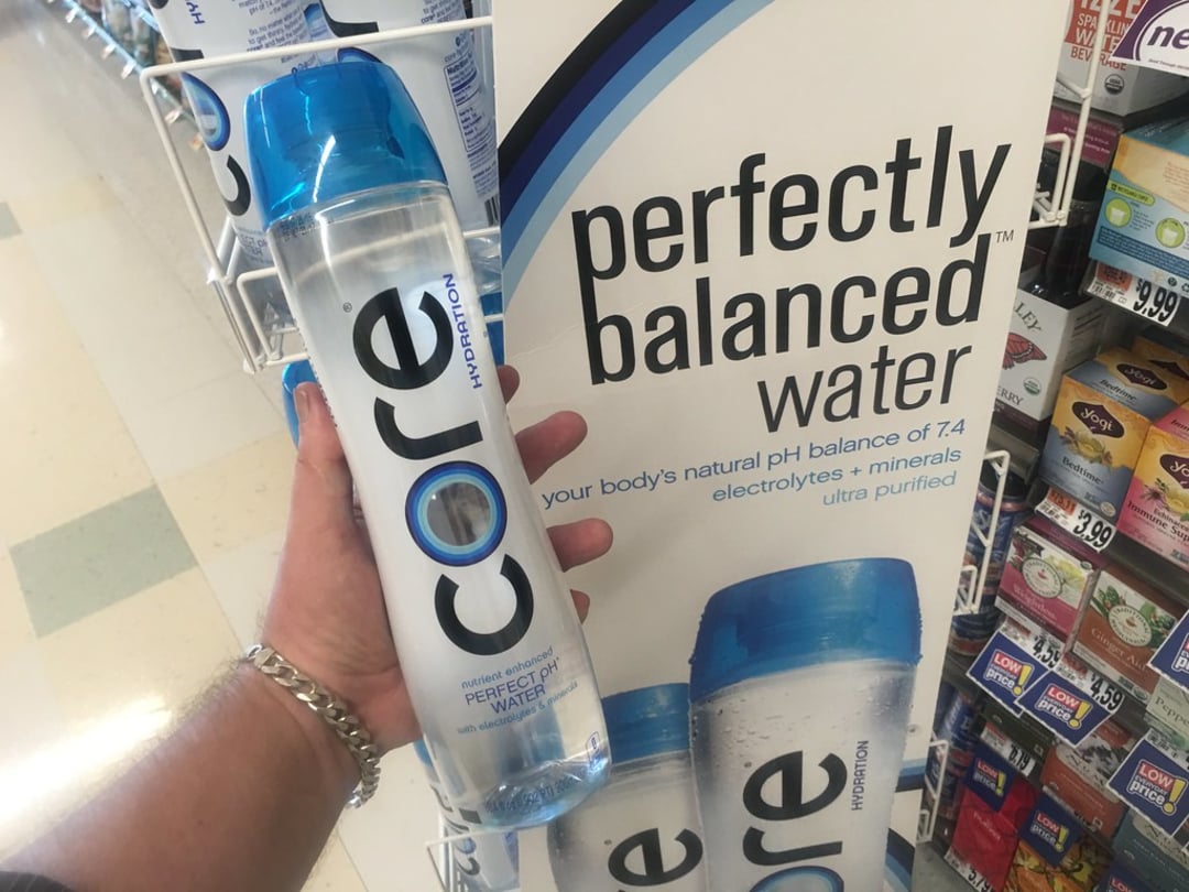 A hand holds a CORE Hydration water bottle in front of a store display sign that reads "perfectly balanced water" and lists its pH balance of 7.4, electrolytes, minerals, and ultra-purified qualities.