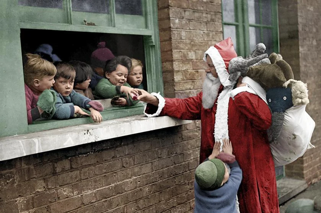 A person dressed as Santa Claus hands treats to children through a window, with more children gathered outside. Santa carries a sack over their shoulder, and a child tugs at Santa’s coat.