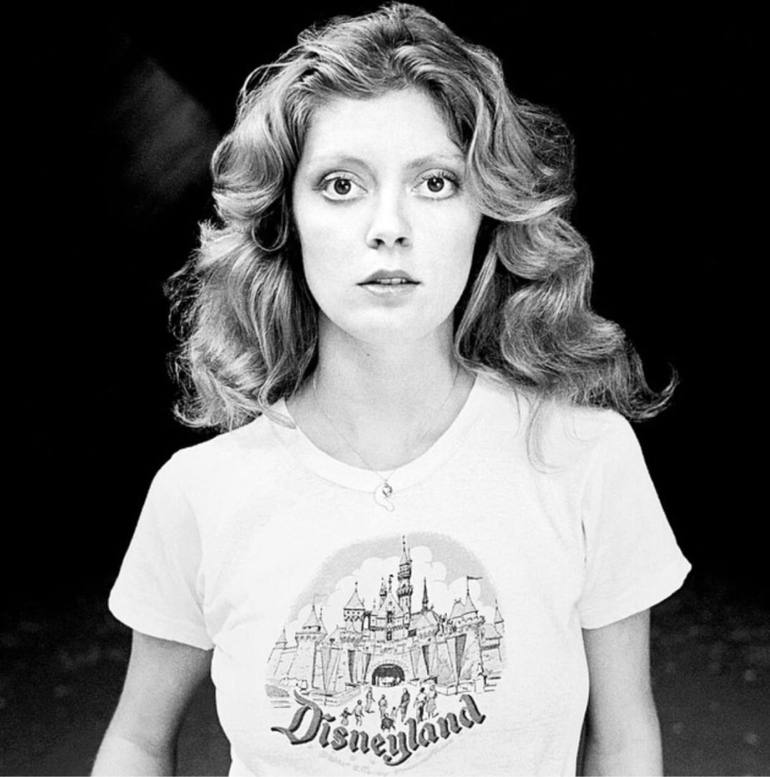 A woman with wavy hair looks directly at the camera, wearing a white T-shirt with a Disneyland graphic and a castle illustration, against a plain dark background.