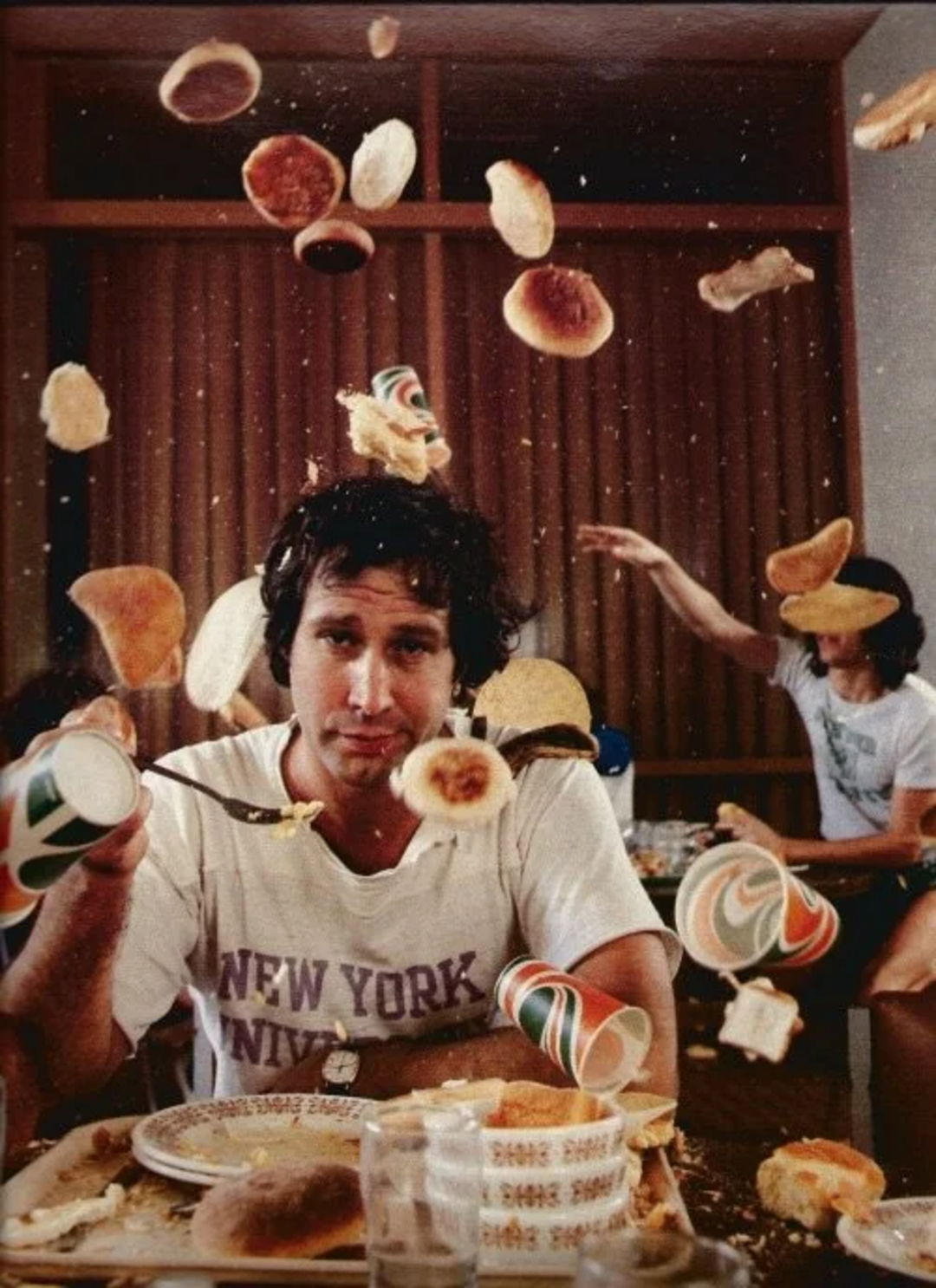 A man in a "New York" t-shirt sits at a table as cups, buns, and food fly through the air around him. Another person in the background is seen throwing items, creating a chaotic, playful scene.