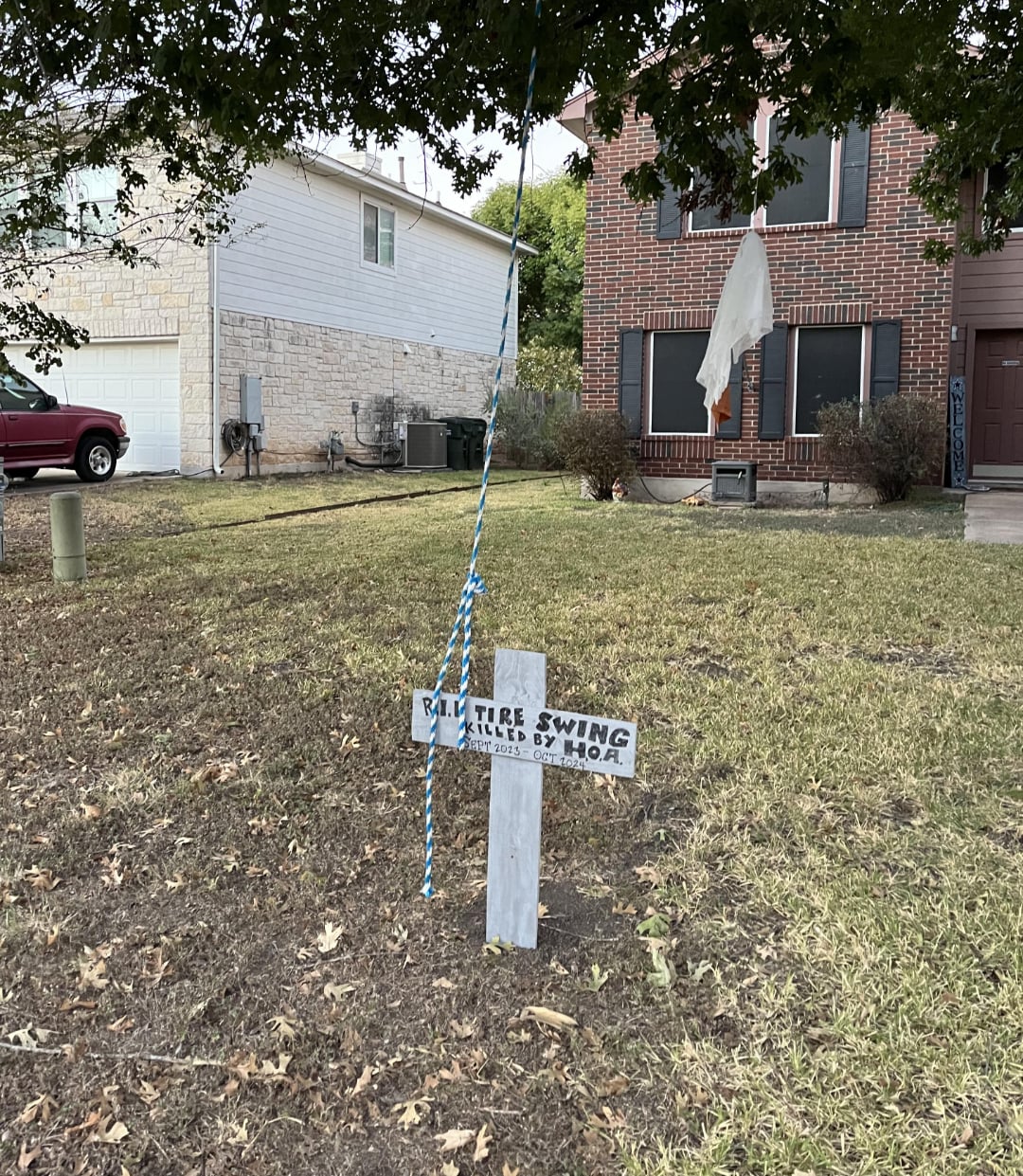 A small wooden sign in a yard reads, "R.I.P. Tree Swing, Killed by HOA." Nearby, a blue rope hangs from a tree branch, indicating where the swing once was. Houses and a ghost decoration are visible in the background.