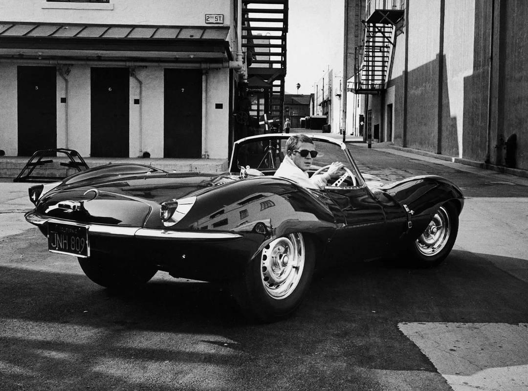 A man wearing sunglasses sits in a sleek, vintage convertible sports car parked on a city street, with buildings and a metal staircase in the background.