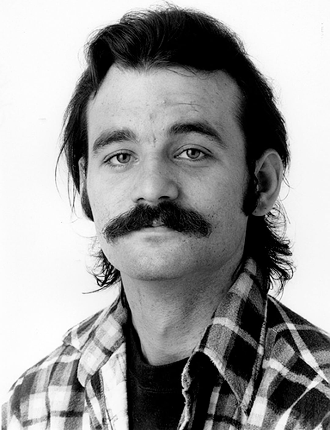 A man with a thick mustache and medium-length dark hair wears a plaid flannel shirt over a dark t-shirt, looking directly at the camera with a neutral expression against a plain white background.