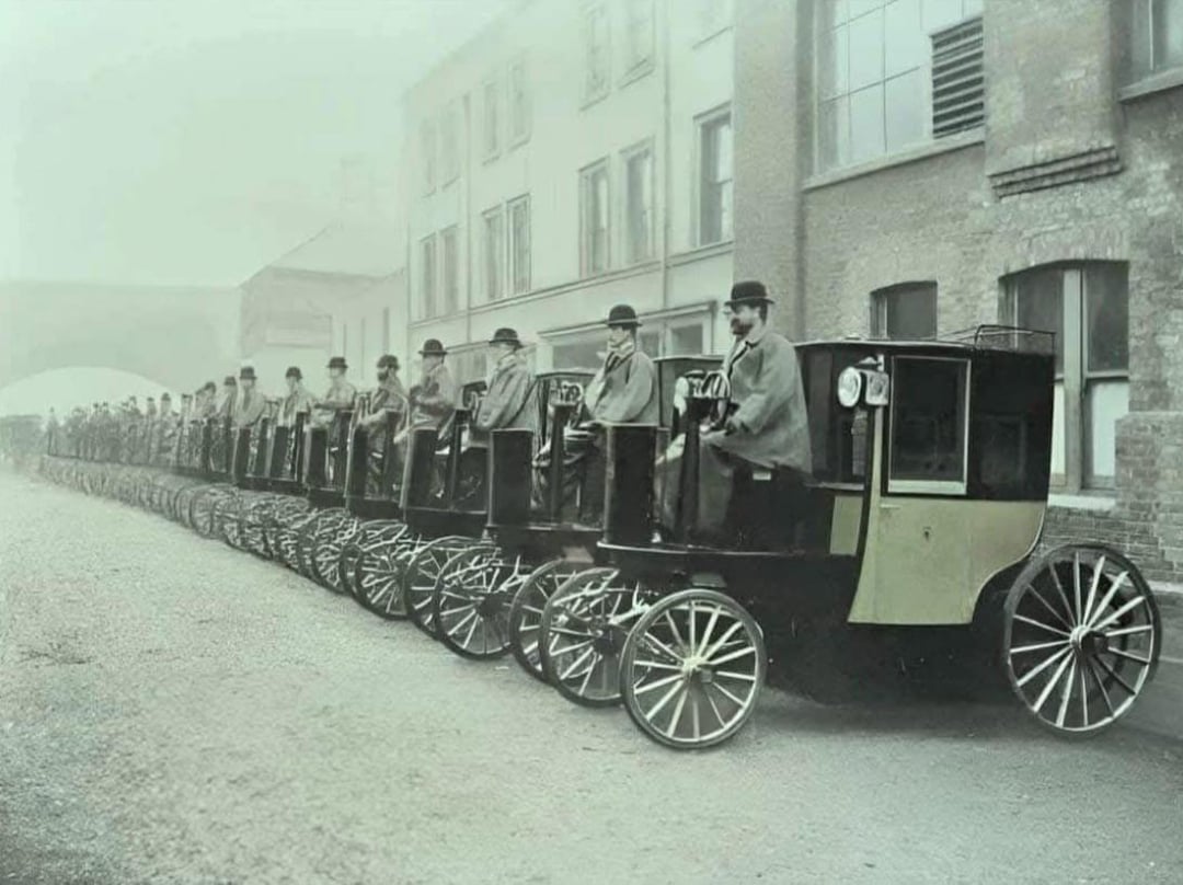 A row of early 20th-century carriages with many wheels is lined up on a street; drivers in hats and cloaks sit in each vehicle, and brick buildings are visible in the background.