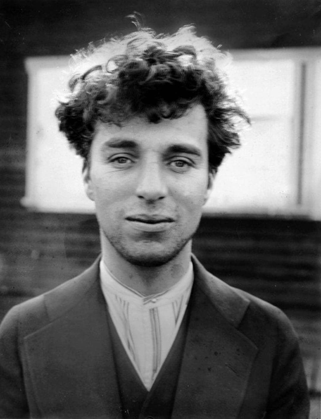 A young man with curly hair and a slight smile wears a suit jacket over a light-colored shirt, standing outdoors with a blurred building and window in the background.