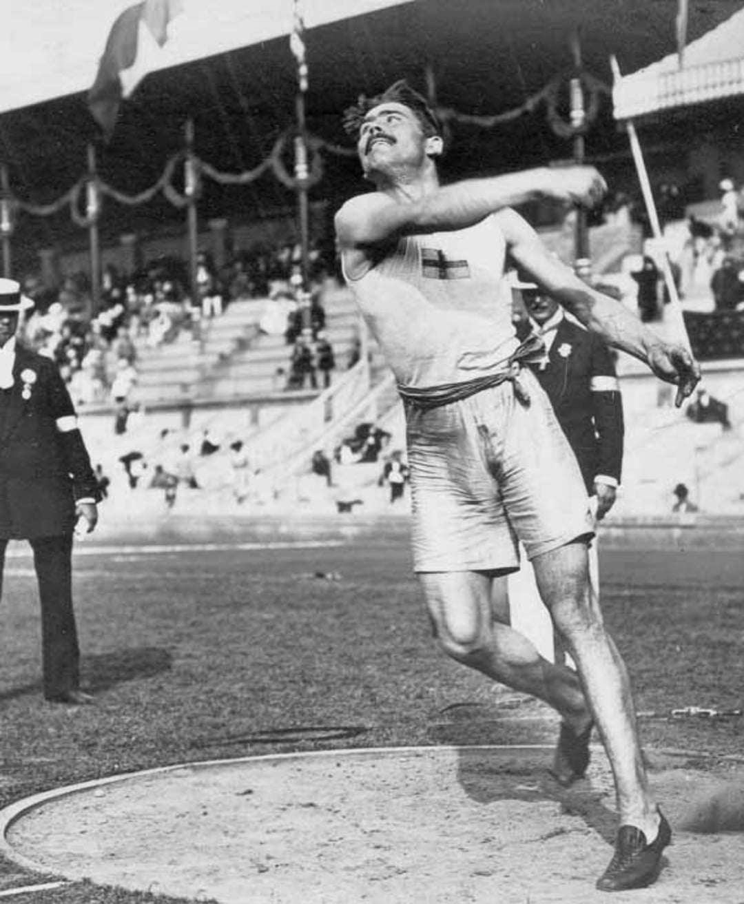 A male athlete in mid-throw during a shot put event at a stadium, wearing athletic shorts and a tank top with a cross emblem. Spectators watch from the stands behind him.