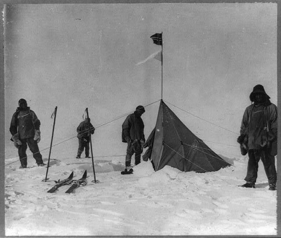 Four people in heavy winter clothing stand beside a tent on a snowy landscape. The tent has a flag on top, and skis and poles are visible in the snow. The sky appears overcast and cold.