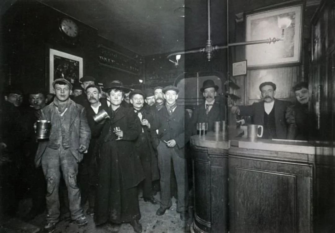 A group of men in old-fashioned clothing gather in a dimly lit pub, some holding mugs. A bartender stands behind the wooden bar. The scene appears to be from the early 20th century.