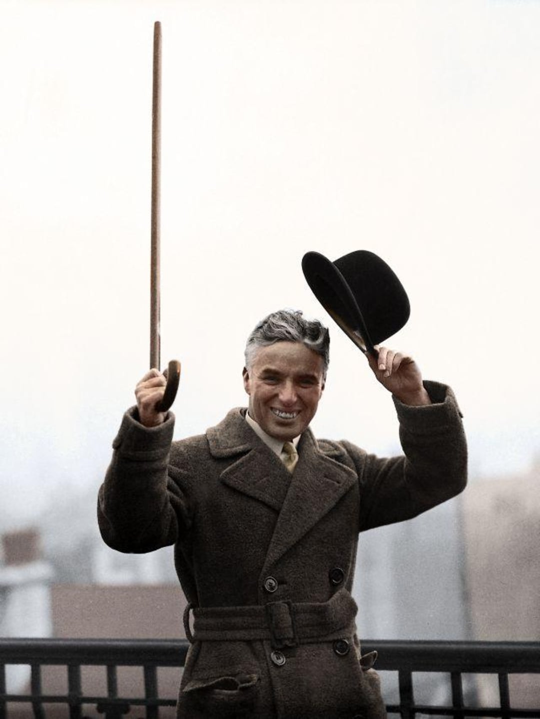 A smiling man in a brown overcoat holds a cane upright in one hand and lifts a black bowler hat with the other, standing outdoors against a blurred background.