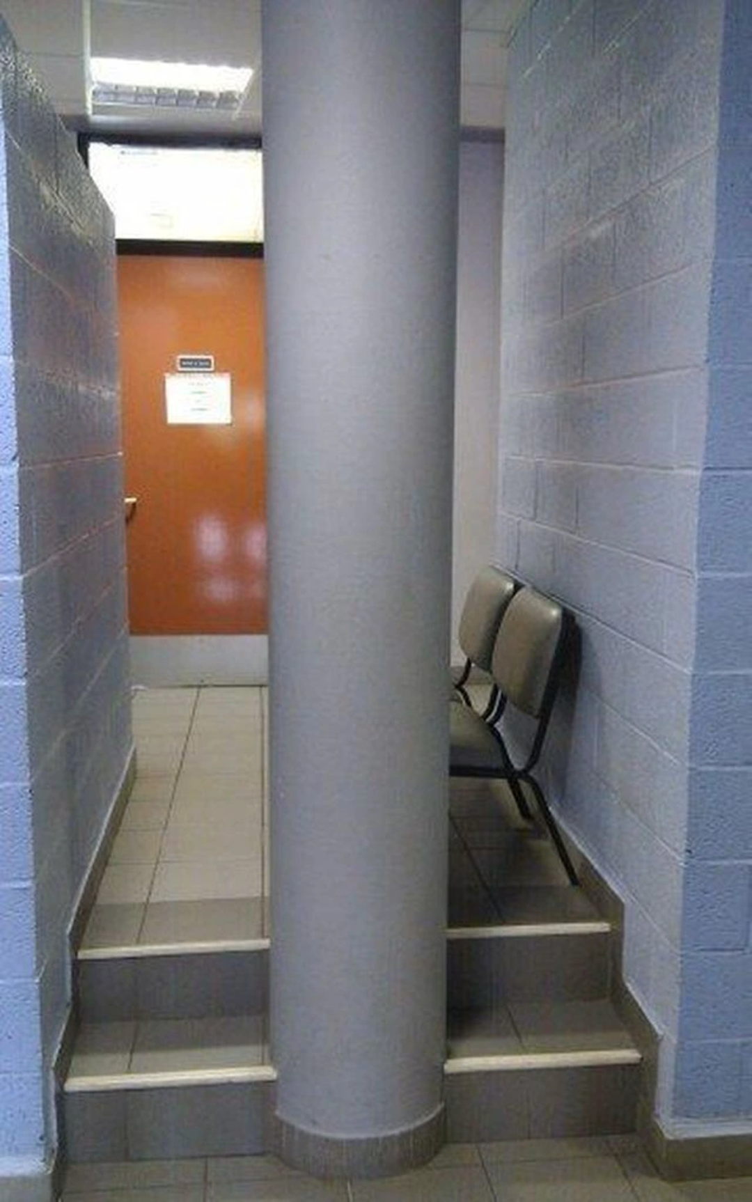 A large column blocks the middle of a narrow hallway, with two chairs on the right and steps leading up to an orange door at the end. The setup makes the hallway and chairs difficult to access.