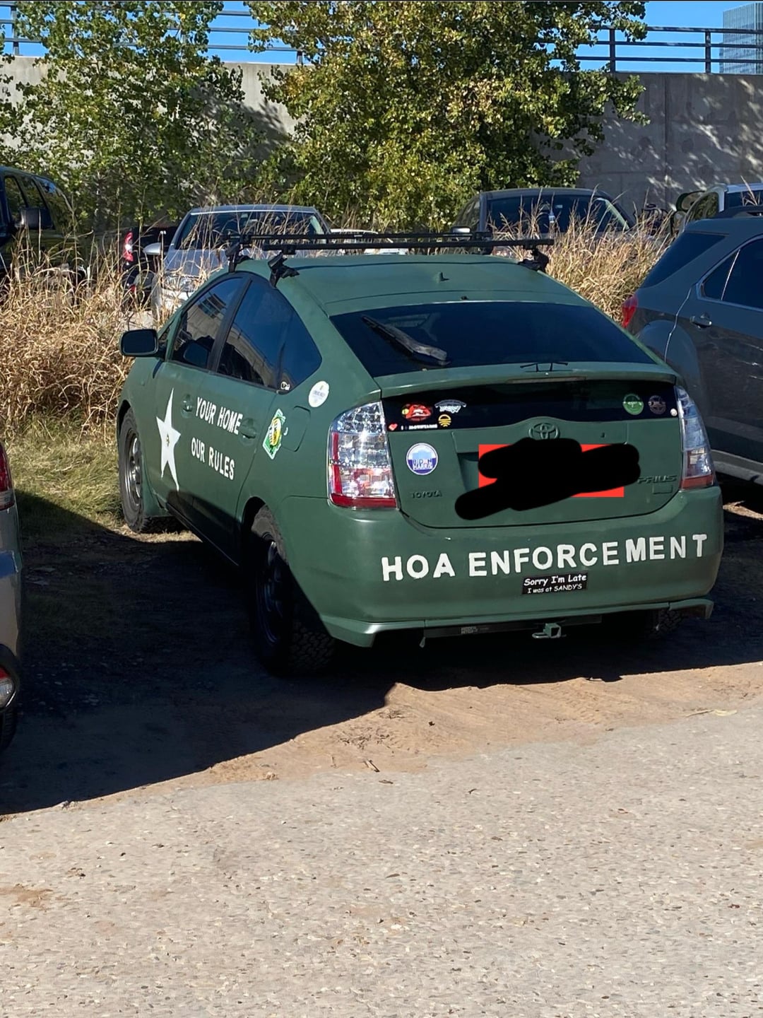 A green Toyota Prius with "HOA ENFORCEMENT" and "Your Home Our Rules" painted on it is parked on grass next to other cars. The vehicle has several bumper stickers and resembles a patrol car.