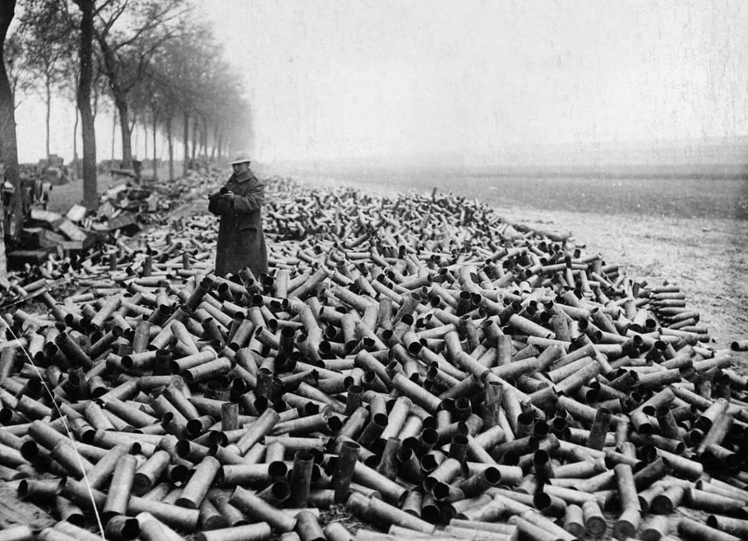 A person in a military coat stands amid a massive pile of empty artillery shell casings on a dirt road lined with trees, in a bleak, foggy landscape.