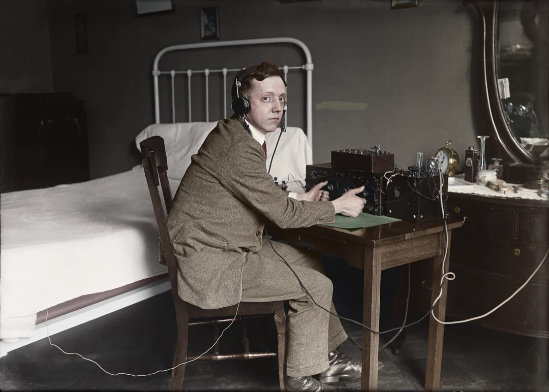 A man in a brown suit sits at a small desk in a bedroom, wearing headphones and operating early radio equipment. A metal bed frame, dresser with bottles, and wall pictures are visible in the background.