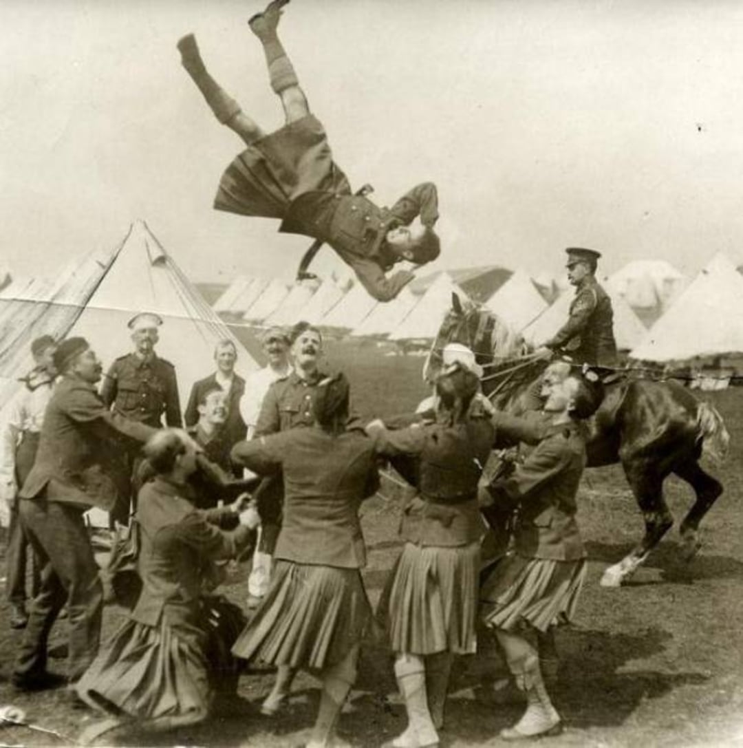 A group of uniformed people toss a man high into the air using a blanket, while others watch. Tents are visible in the background, and a man on horseback observes the scene.