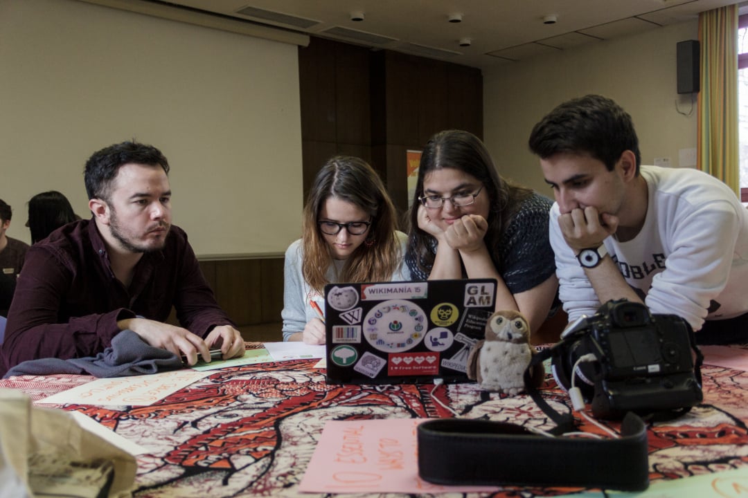 Four young adults sit around a table, intently looking at a laptop covered in stickers. Papers, a camera, and a small toy owl are on the table. The setting appears to be a casual meeting or study session indoors.