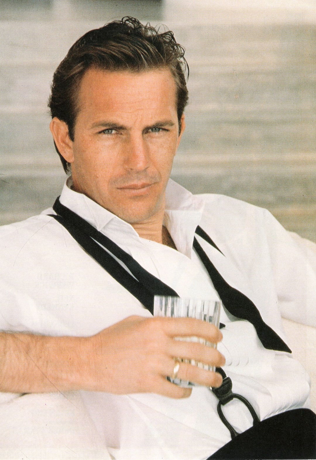 A man with short brown hair wearing a white shirt and a black tie draped around his neck sits relaxed, holding a glass of clear liquid and looking directly at the camera with a serious expression.