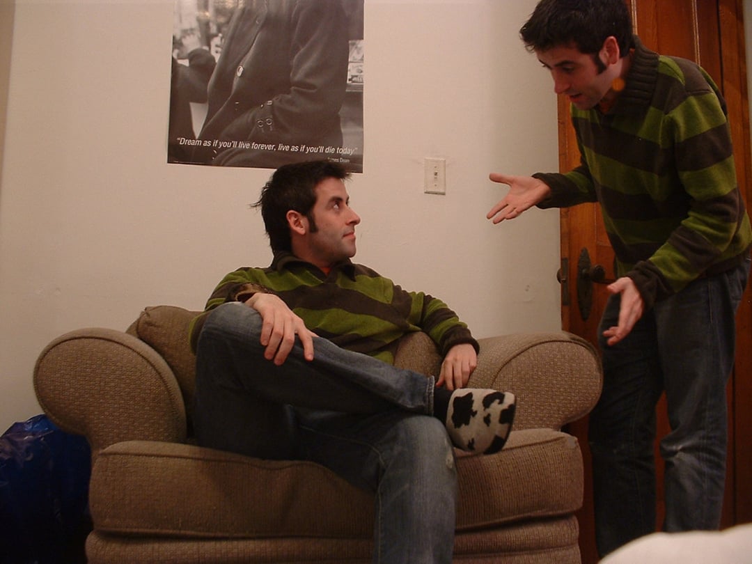 Two men wearing matching green-striped sweaters, who appear identical, are in a room. One sits relaxed on an armchair with crossed legs, while the other stands nearby gesturing animatedly with both hands.