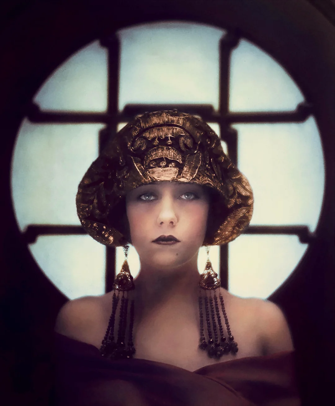 A woman in a dramatic headpiece and long earrings poses in front of a circular, window-like background. She wears dark makeup, a bare-shouldered gown, and gazes directly at the camera with a mysterious expression.