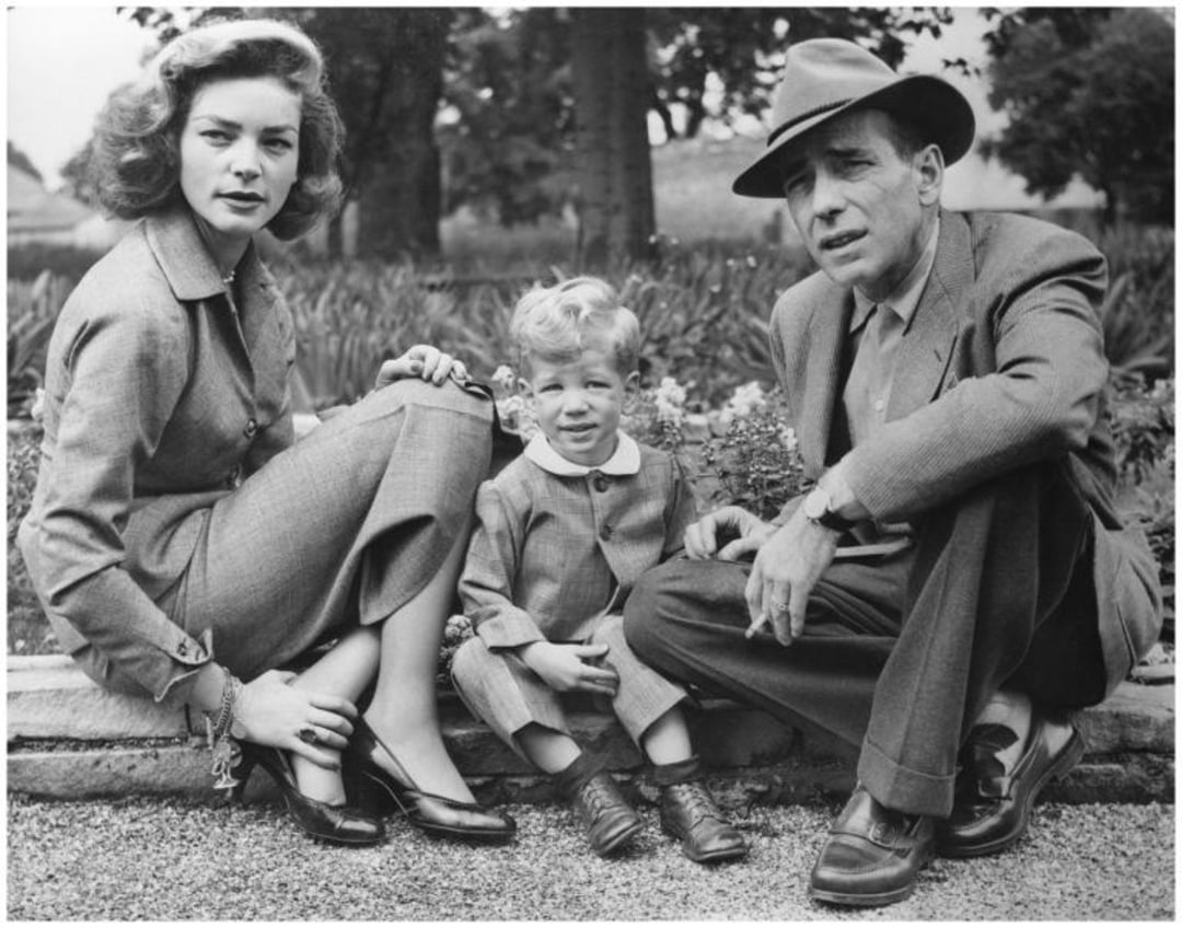 A woman, a young boy, and a man sit outdoors on a path. All wear suits; the woman has wavy hair and heels, and the man wears a fedora. Behind them are trees and greenery. They look toward the camera.