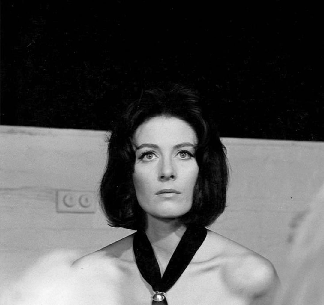 Black and white photo of a woman with dark, shoulder-length hair, wearing a halter-style top or necklace, looking slightly upward with a neutral expression. The background is plain with a power outlet visible on the wall.