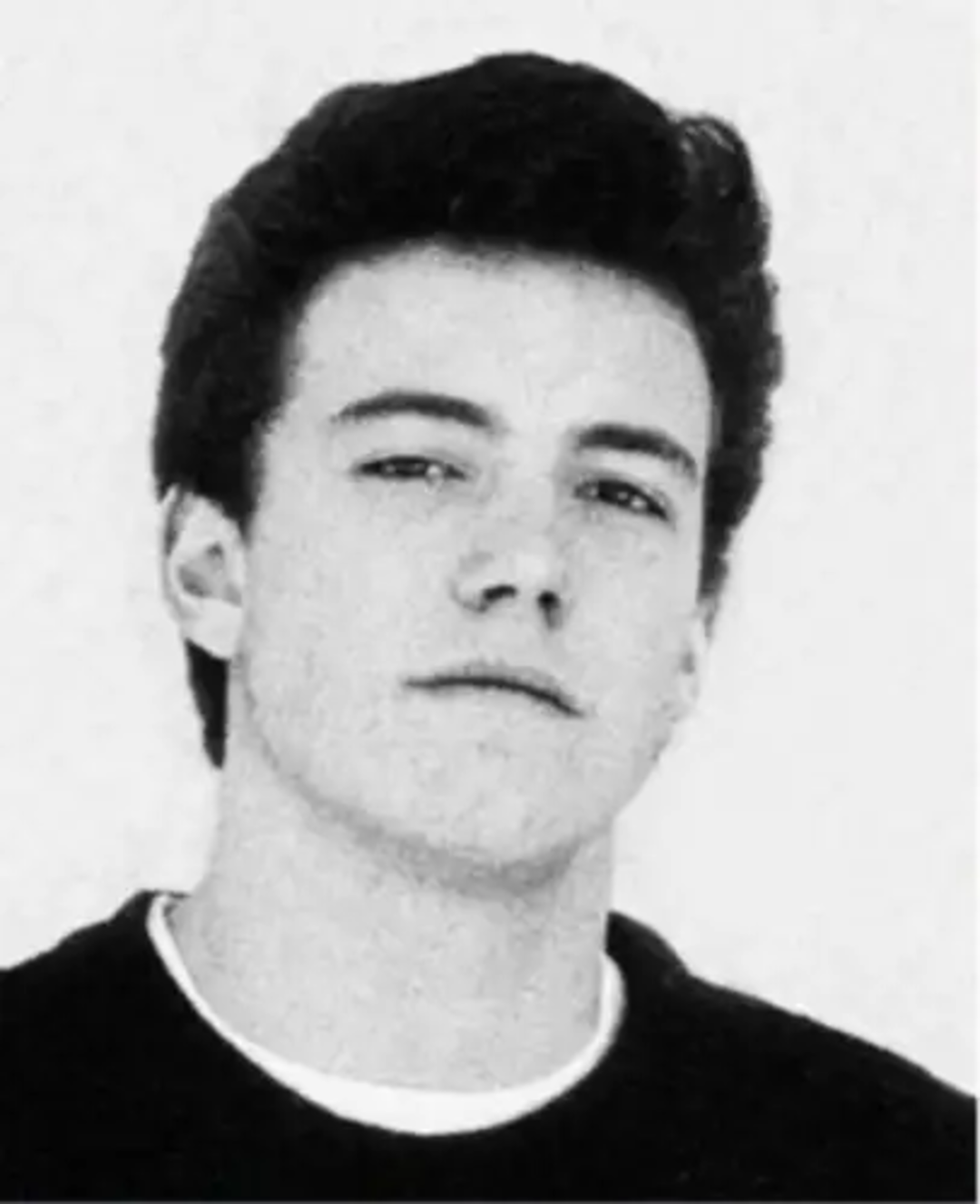 Black and white photo of a young man with short, dark hair, wearing a dark shirt over a white crewneck. He is facing forward with a neutral expression against a plain, light background.