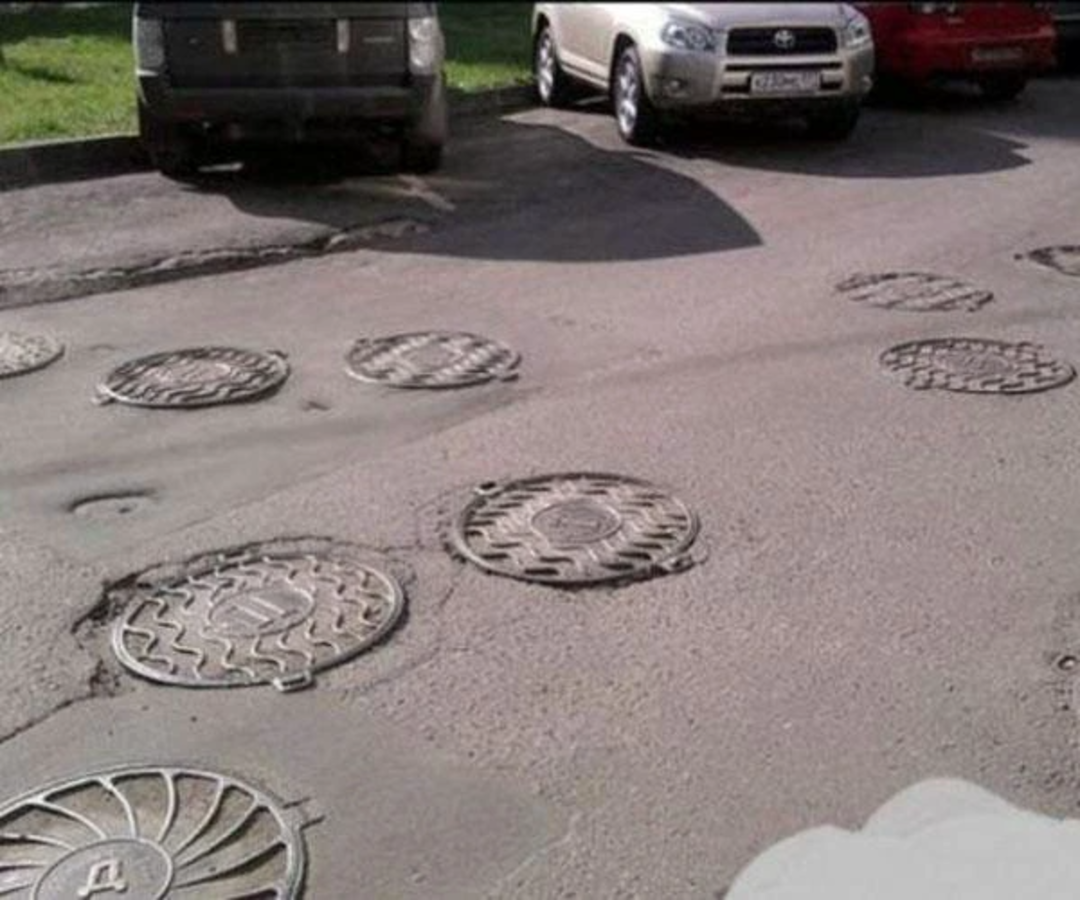 Several manhole covers are scattered unevenly across a damaged asphalt road in a parking lot, with parked cars visible in the background. The placement of the covers appears random and unusual.