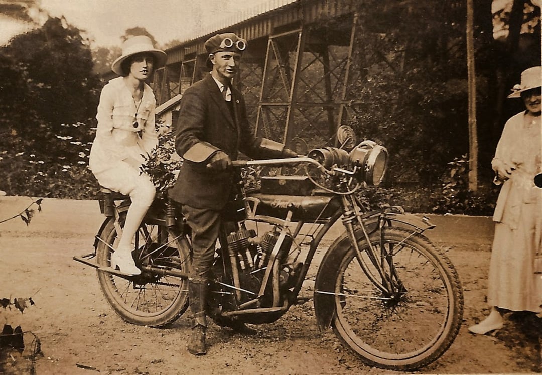 A vintage photo of a man in a suit and goggles standing beside an old motorcycle, with a woman in a dress and hat sitting on the back. Another woman in a hat stands nearby, and a metal bridge is in the background.