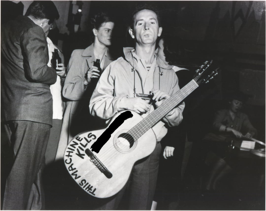 Black and white photo of a man standing and holding a guitar with the words "THIS MACHINE KILLS" written on it; people are talking and sitting in the background at what appears to be a social gathering.