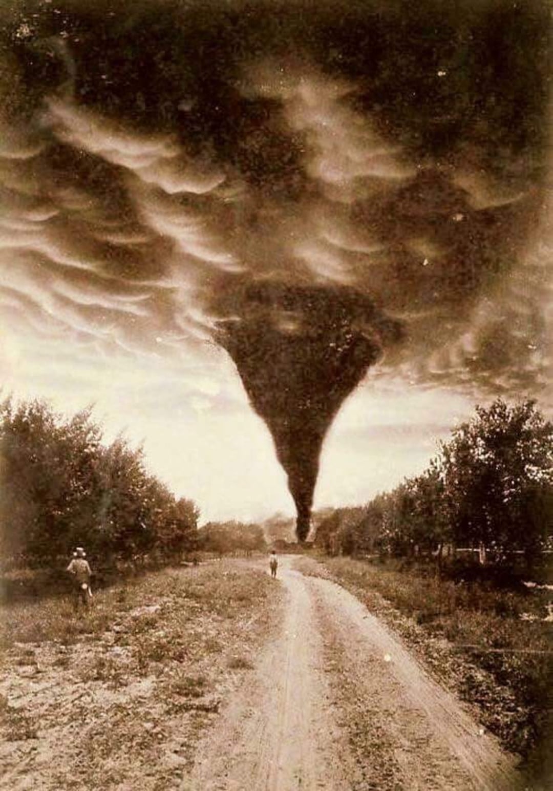 A sepia-toned photo shows a powerful tornado touching down in the distance, centered on a dirt road lined with trees. Two people are seen on the road, one closer to the tornado and another to the left side.